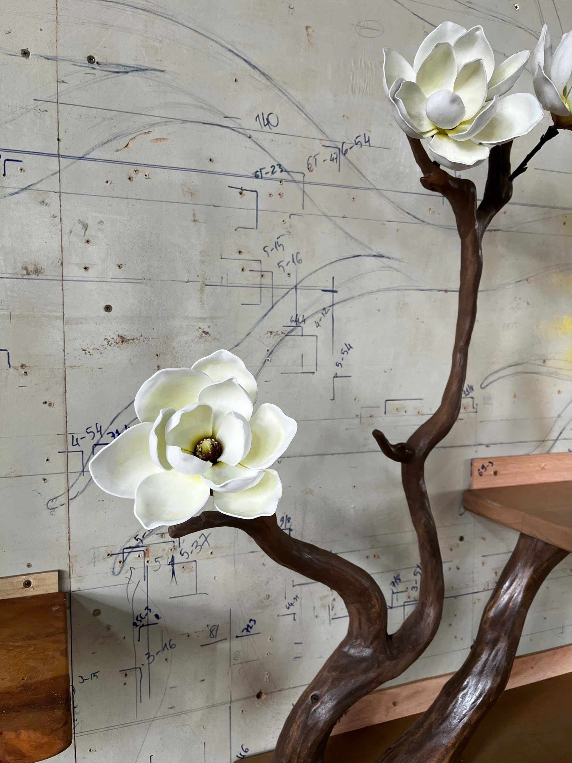 Decorative plant with white flowers on a wooden stand against a wall with blueprints.