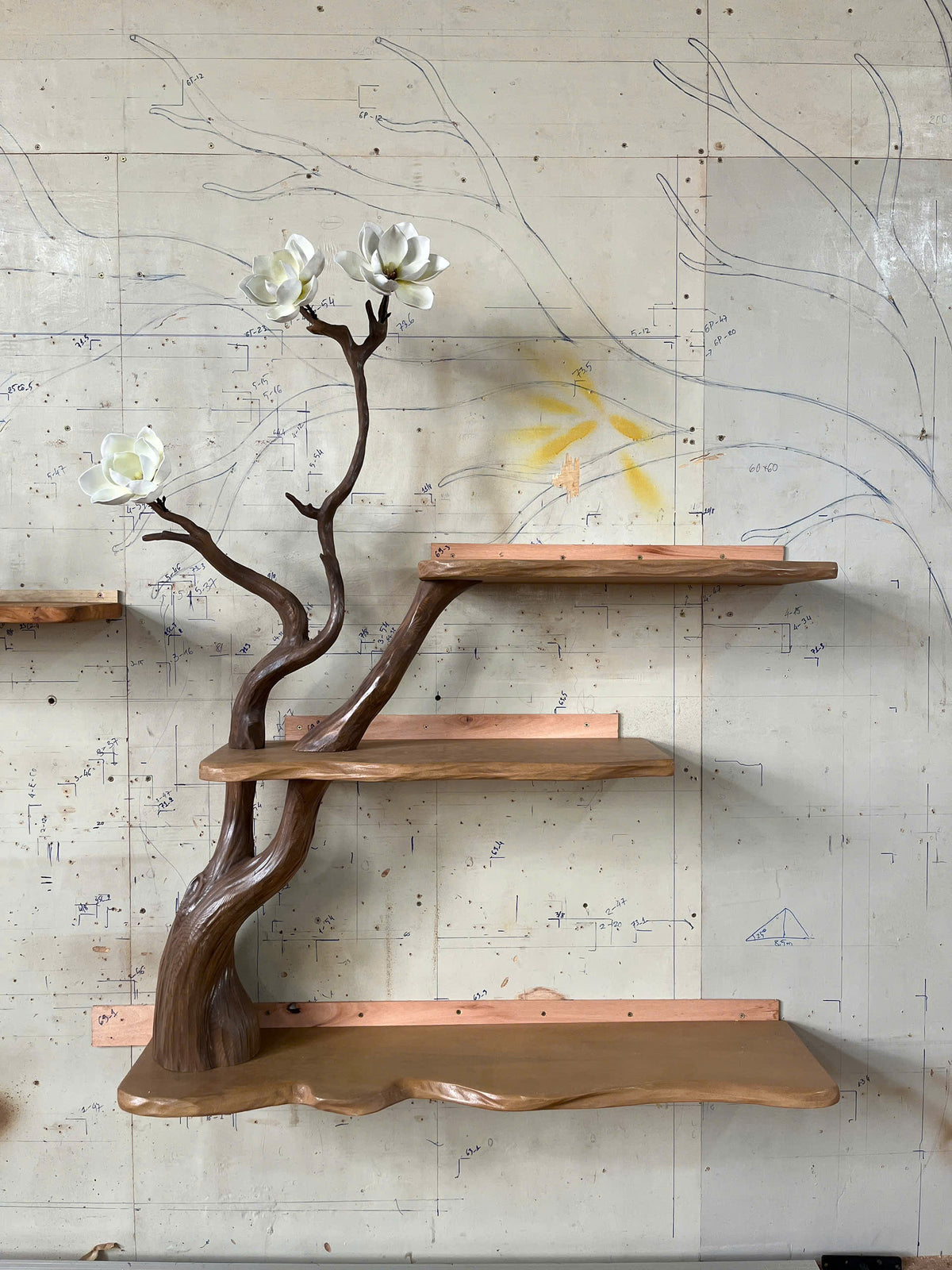 Wooden shelf designed like a tree branch with white flowers against a textured wall.